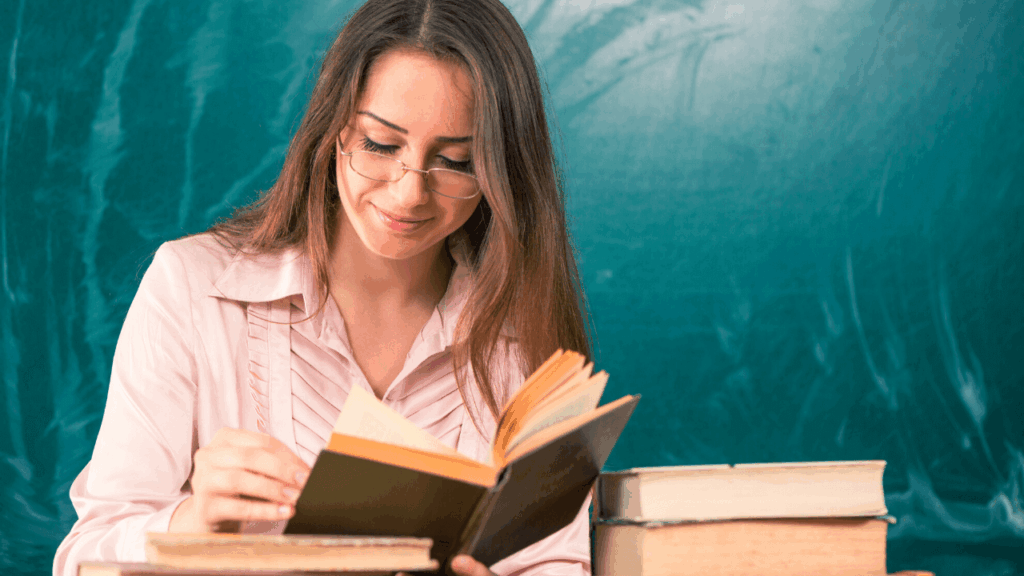 Mulher lê um livro. Na mesa à frente dela tem uma pilha de outros livros fechados.